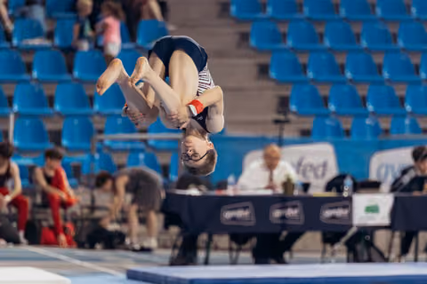 Young gymnast performs an inverted tucked flip mid-air, focused expression, blue arena seating in background.