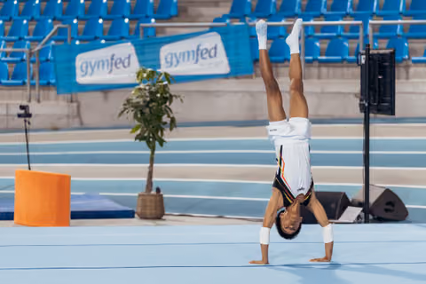 Young gymnast performing a handstand on floor exercise mat in an indoor arena with blue seating and gymfed banners.