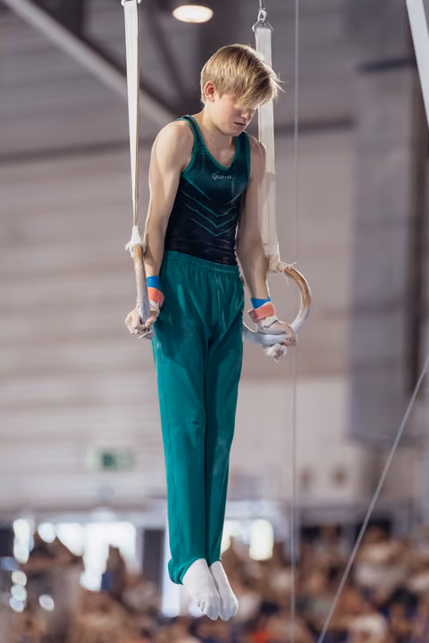 Young male gymnast hangs focused on still rings, wearing green uniform and grip guards, in a busy sports hall.