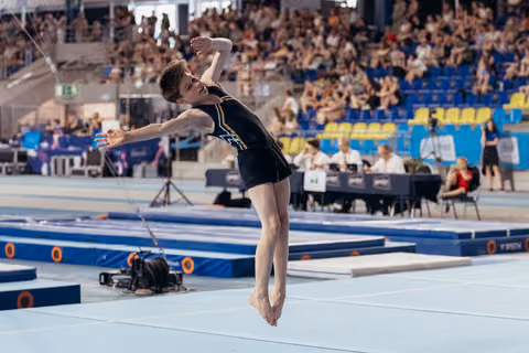Young male gymnast soars mid-air during floor routine, arms outstretched, focused expression, packed arena in background.