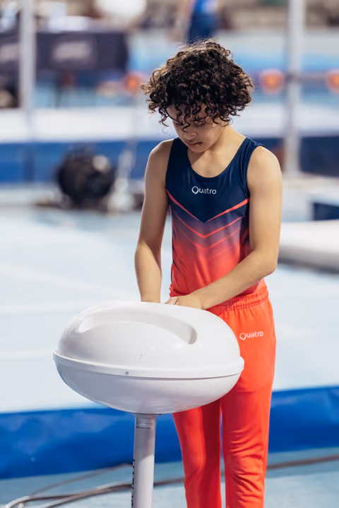 Young gymnast with curly hair focuses intently while gripping a chalk bowl, wearing a Quatro leotard at a competition venue.