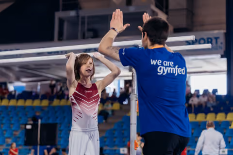 Young gymnast in red leotard high-fives her coach after uneven bars routine, both arms raised in celebration.