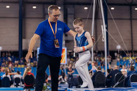 Coach in blue shirt steadies a focused young gymnast on the trampoline during a gymnastics competition.