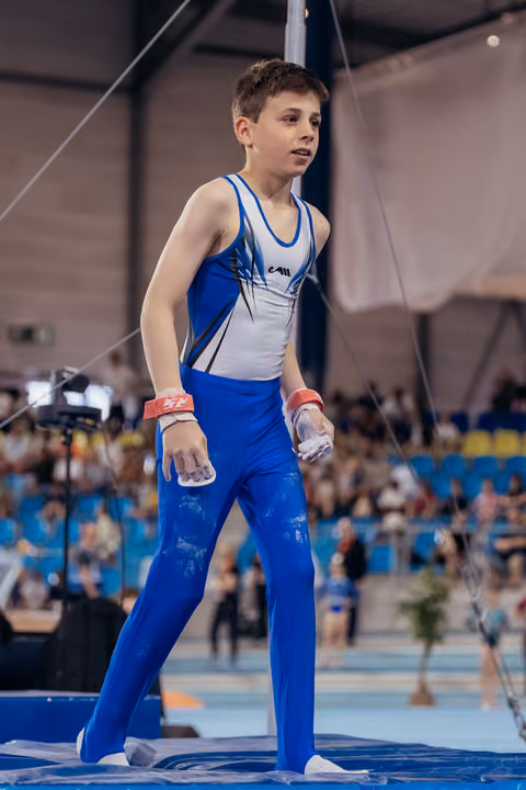 Young male gymnast stands poised on mat, chalk-covered hands, looking aside with calm focus during indoor gymnastics event.