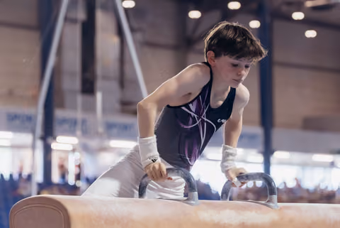 Young gymnast concentrates intensely while performing on the pommel horse in a busy gymnastics hall.
