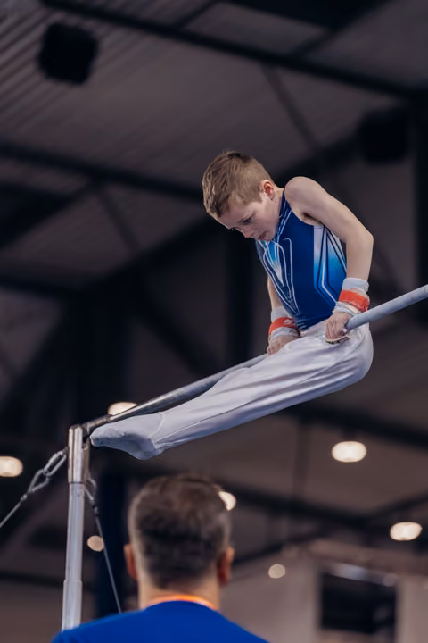 Young boy gymnast grips the high bar with intense focus, legs extended, wearing blue leotard and wrist guards indoors.