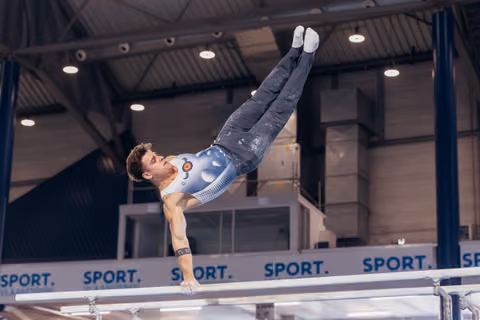Male gymnast performs a dramatic horizontal hold on parallel bars, body fully extended, intense focus in a sports arena.