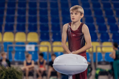 Blonde boy gymnast in dark red leotard stands at chalk bowl, gazing intently aside in a competition arena.