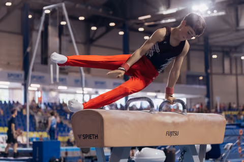 Young male gymnast performs scissor move on pommel horse, leaning forward with focused intensity in packed arena.