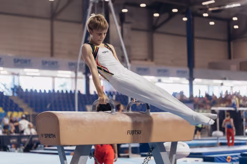 Young male gymnast performs a focused pommel horse routine in a competitive gymnastics hall, wearing a white and black leotard.