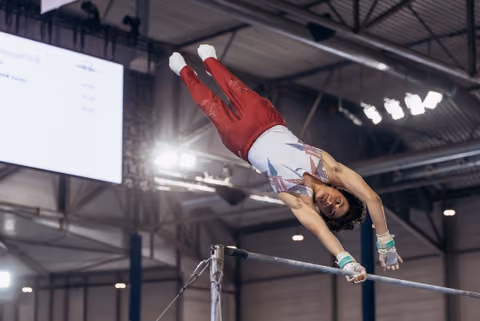 Male gymnast releases the high bar mid-flight, body fully extended and inverted, competing in an indoor gymnastics arena.