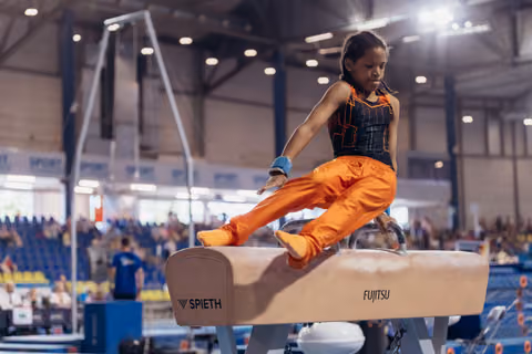 Young gymnast performs on the pommel horse with intense focus, wearing an orange uniform in a packed arena.