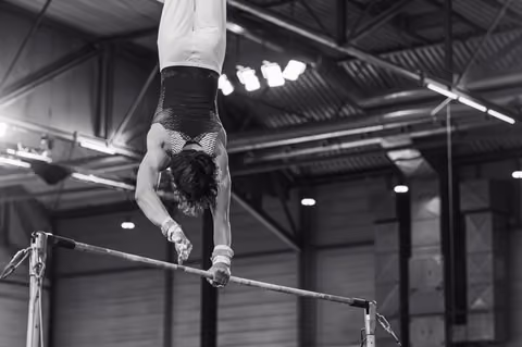 Male gymnast performs an inverted handstand on the high bar, muscles tensed, in a large indoor gymnastics hall.