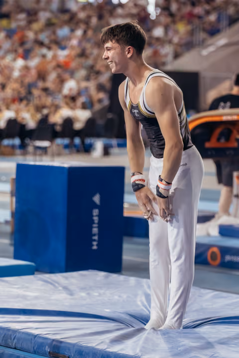 Young male gymnast smiling confidently, chalked hands clasped, standing on mat beside blue Spieth vault block in packed arena.