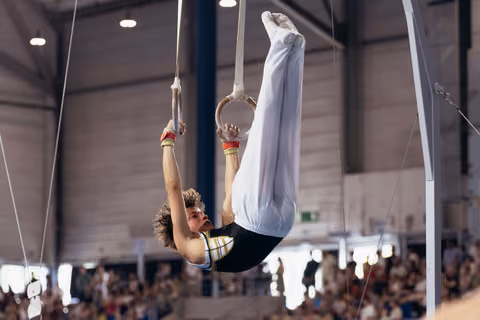 Young male gymnast performs an inverted pike hold on still rings, showing intense focus and strength before a crowd.