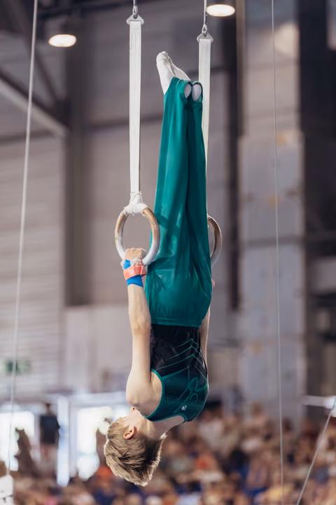 Young male gymnast performs an inverted hold on still rings, body fully extended, in a packed competition hall.