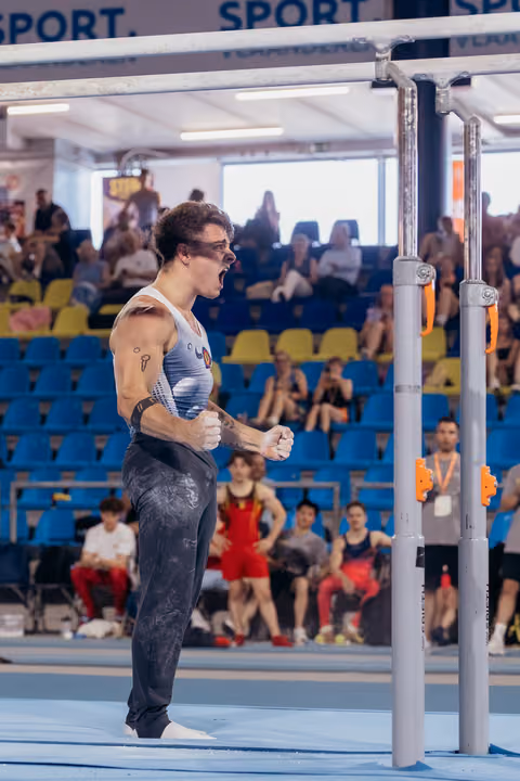 Male gymnast celebrates with fists clenched and mouth open in triumph beside parallel bars, crowd watching in background.
