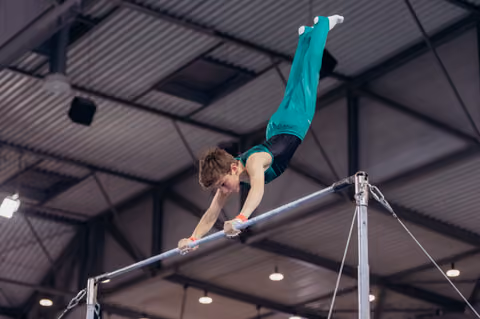Young male gymnast grips the high bar with fierce concentration, body extended horizontally in a dynamic gymnastics hall.