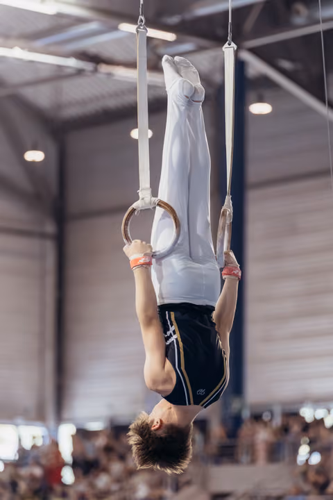 Young male gymnast performs an inverted hang on still rings, wearing white and navy, in a packed indoor arena.
