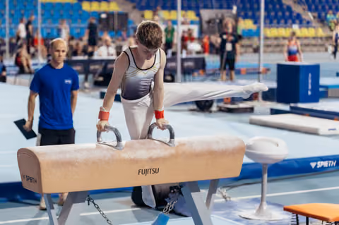 Young male gymnast performs on the Fujitsu pommel horse, legs extended, concentrating intensely during a competition.