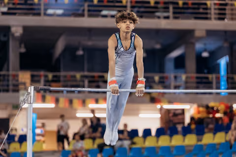 Young male gymnast grips the high bar with focused intensity, body extended, wearing red wrist guards in a sports arena.