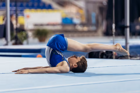 Young male gymnast performs a bridge pose on floor exercise mat, upper body stretched flat, legs extended upward.