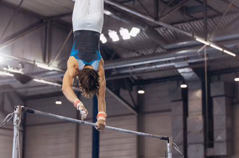 Male gymnast performs an inverted handstand on the high bar, muscles tensed, in a large indoor sports hall.