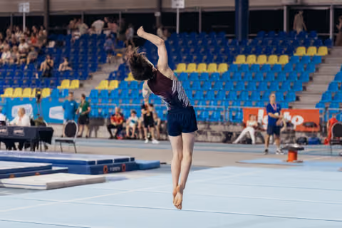 Young gymnast mid-air on floor exercise, one arm raised overhead, poised and focused during indoor competition.