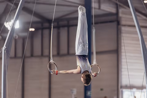 Young male gymnast performs a perfect inverted hang on still rings, body fully extended, in an indoor gymnasium.