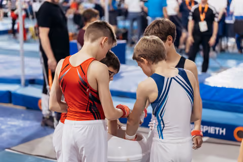 Young male gymnasts help each other wrap wrists before competing, showing camaraderie in a busy gymnastics hall.