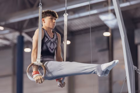 Young male gymnast holds a straddle position on still rings, expression focused and determined, in an indoor gymnasium.