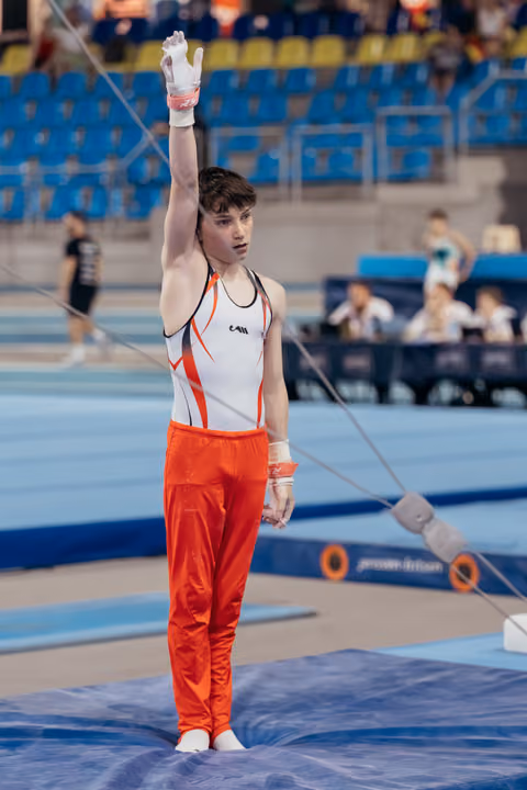 Young male gymnast raises one arm in salute, composed expression, wearing orange and white leotard on competition mat.