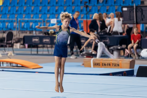 Young male gymnast leaps mid-air on floor mat, arms outstretched, focused expression during indoor gymnastics event.