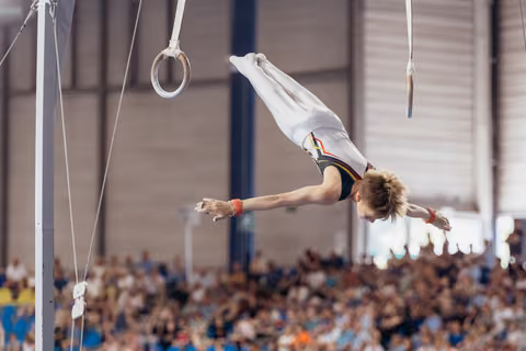 Young male gymnast holds a dramatic horizontal position on still rings, arms extended, body parallel to the floor in an indoor arena.