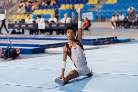 Young male gymnast sits on floor mat, one arm raised high in a composed finishing pose during competition.