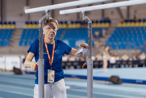 Older Belgian official stands focused between parallel bars in an indoor athletics stadium, holding a paper.