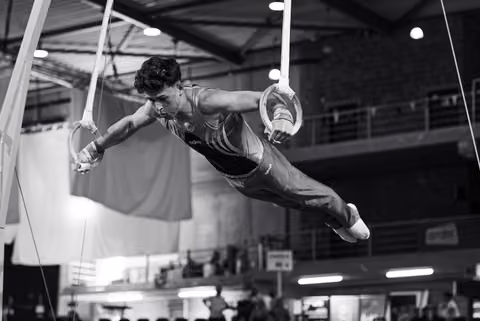 Young male gymnast holds an iron cross on still rings, body horizontal, expression intense with focused determination in indoor arena.