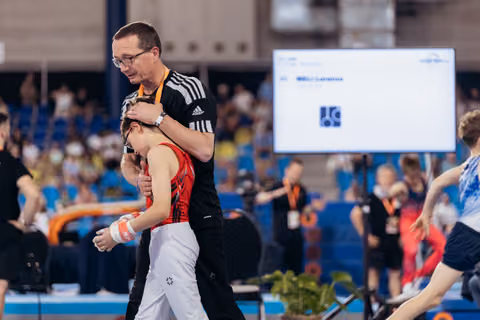 A coach embraces a young gymnast in a red leotard, offering comfort after a performance in a busy arena.