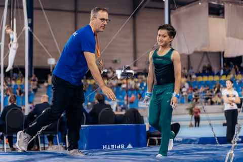 Coach in blue leans toward young gymnast in green uniform, sharing a warm interaction on the trampoline floor at indoor arena.