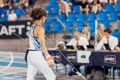 Young male gymnast stands with back to camera, facing judges at Gymfed table, composed before or after apparatus.