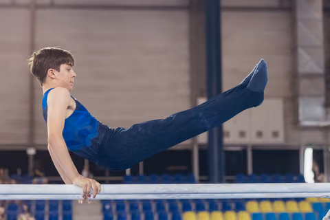 Young male gymnast performs a straddle L-sit on parallel bars, focused expression, in an indoor sports arena.