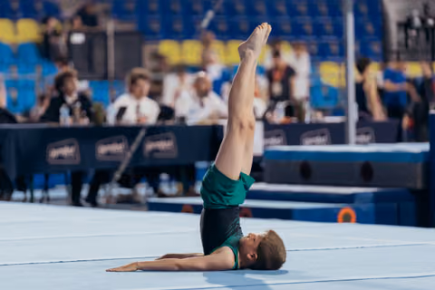 Young gymnast performs a shoulder stand on the floor exercise mat, legs extended vertically, during a competition.