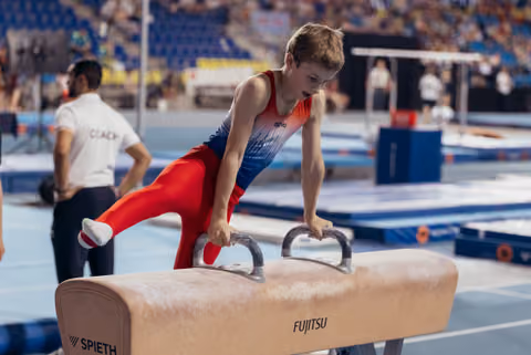 Young male gymnast performs on pommel horse with intense concentration, mouth open, leaning forward at indoor gymnastics event.