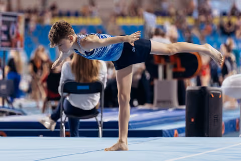 Young male gymnast holds a precise arabesque balance on floor, arms extended, expression focused and determined.