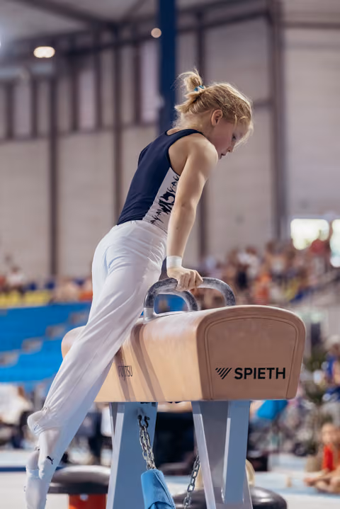Young male gymnast grips pommel horse handles with intense focus, leaning forward mid-routine at indoor competition.