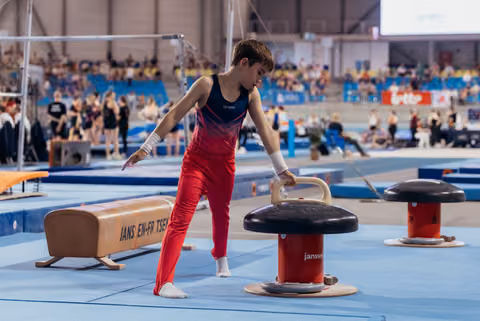 Young male gymnast in red leans intently over pommel horse, adjusting grip with focused concentration at indoor competition.