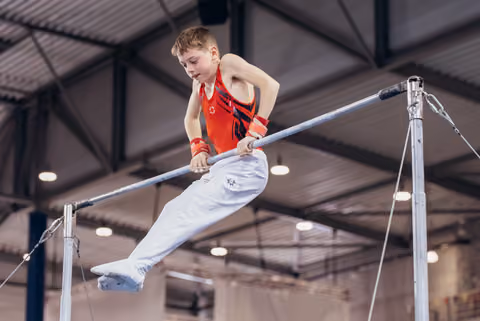 Young male gymnast grips the high bar with intense focus, legs extended mid-routine in an indoor gymnastics hall.