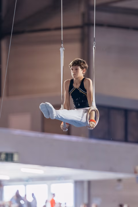 Young male gymnast holds a tucked seat position on still rings, focused expression, indoor gymnastics hall.