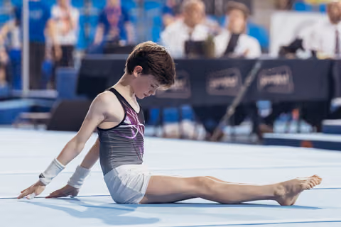 Young gymnast sits in a straddle stretch on the floor mat, head bowed in concentration during a competition.
