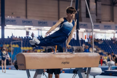 Young male gymnast performs a focused pommel horse routine, body suspended mid-swing in a packed sports hall.